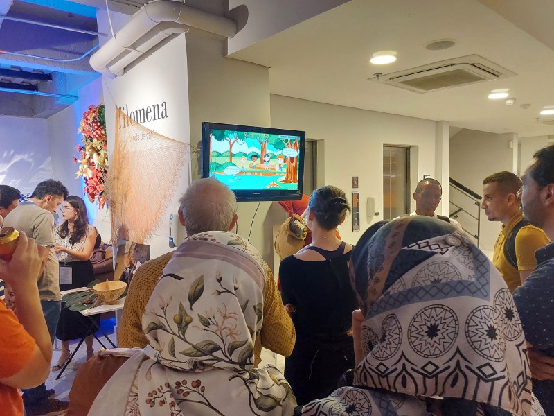 A large mixed group of people gather around a screen to watch videos from Hub members during the ECR Showcase event